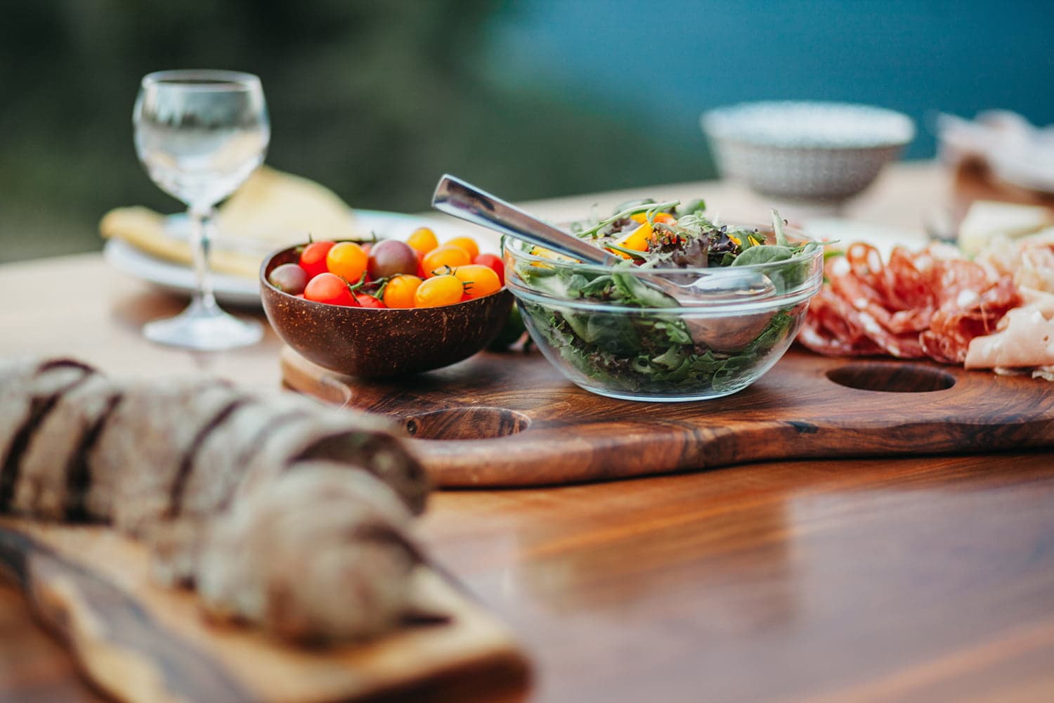 A wooden platter holds a salad, cherry tomatoes, and assorted meats, with a glass of wine in the background.