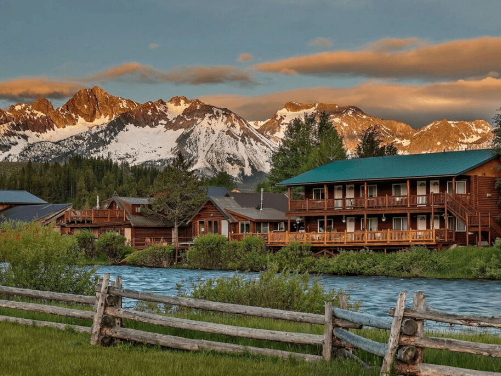 Lodges sit beside a river with snowy mountains in the background under a colorful sky.