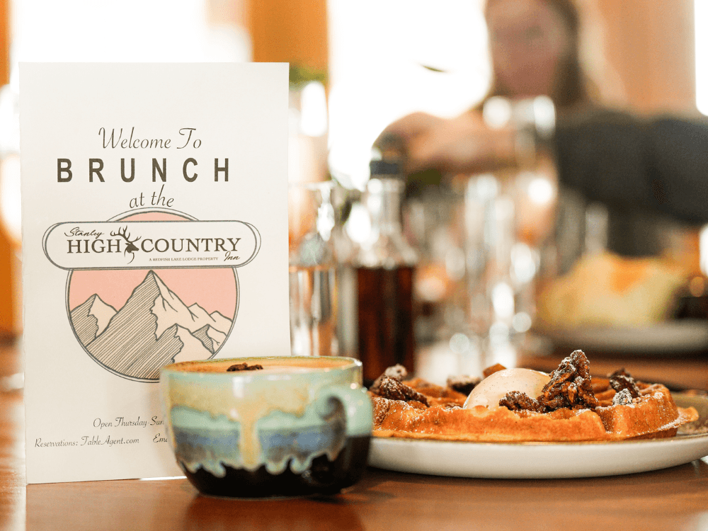 A brunch menu stands next to a plate of waffles and a coffee cup on a wooden table.