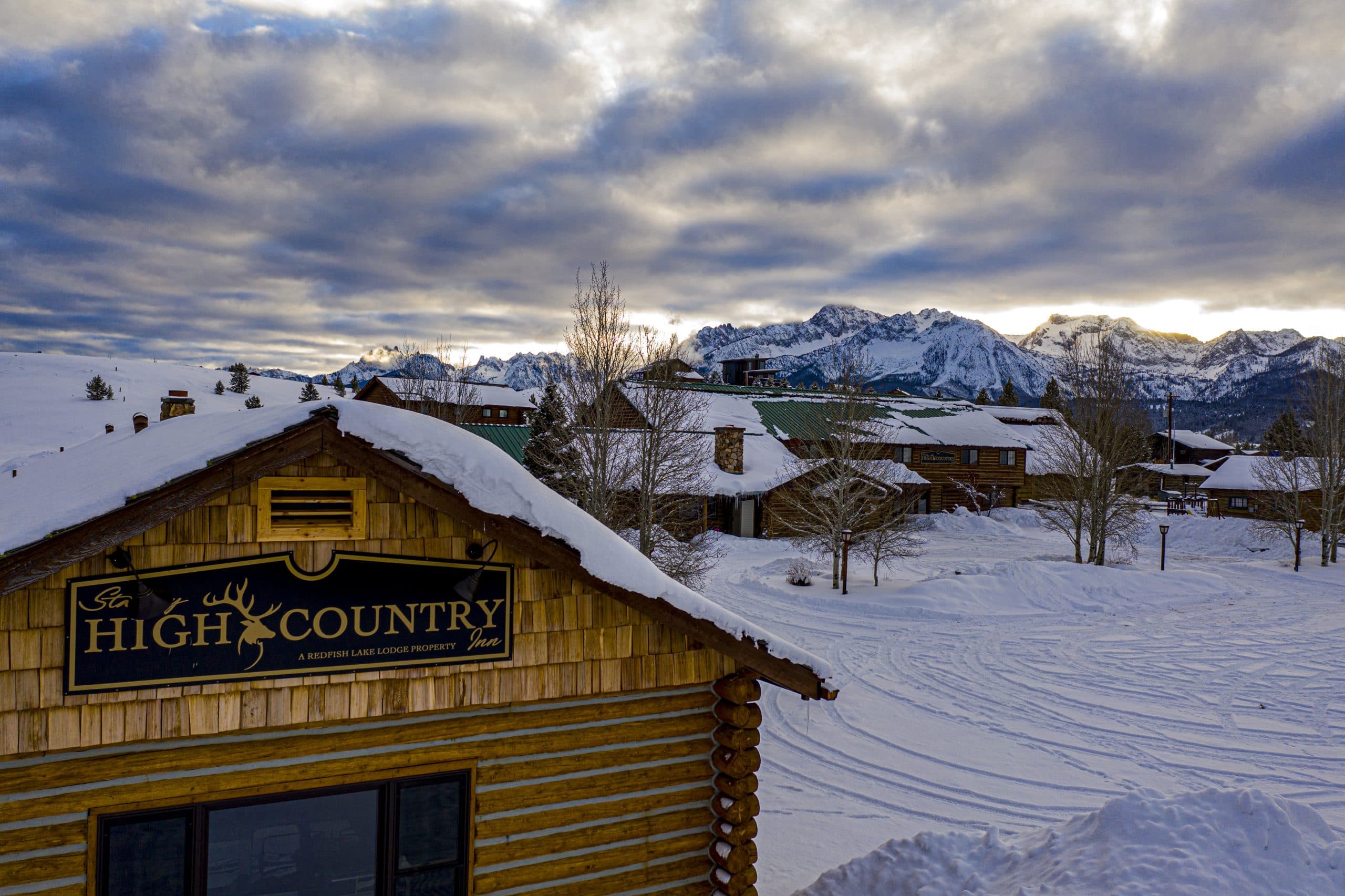 A log cabin with the sign "High Country" surrounded by snowy landscape and mountains under a cloudy sky. A log cabin with the sign "High Country" surrounded by snowy landscape and mountains under a cloudy sky.
