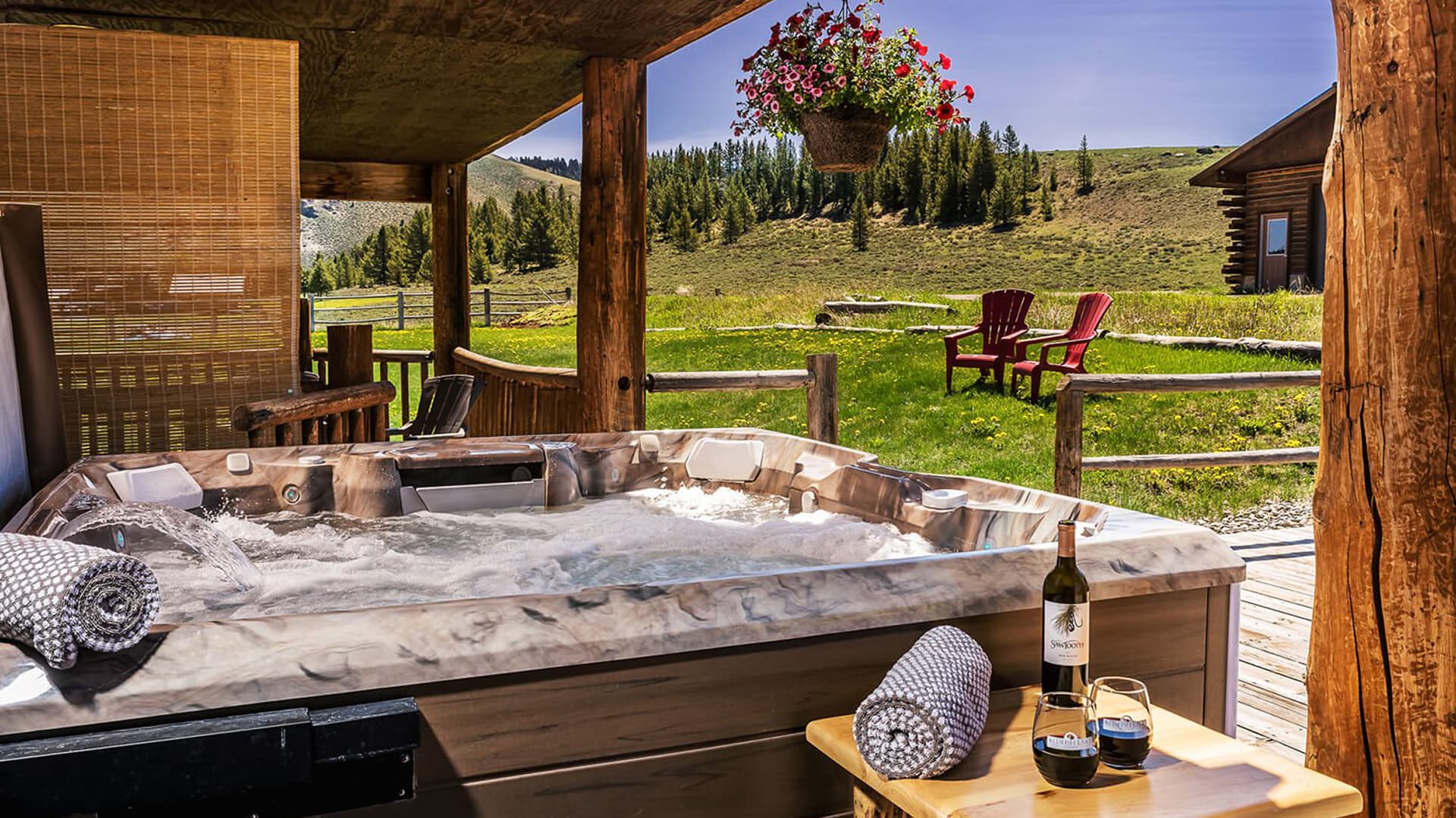 A hot tub on a wooden deck overlooks a scenic landscape with chairs and a bottle of wine nearby.