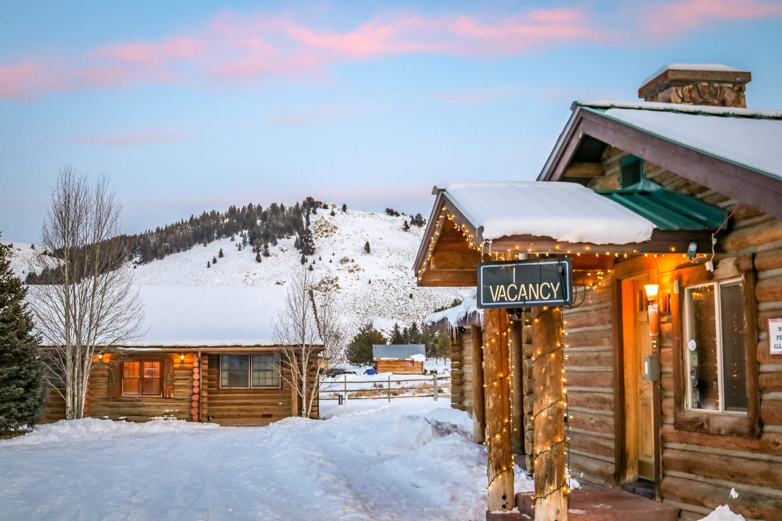 A cozy log cabin with a "VACANCY" sign surrounded by snow-covered hills at dusk. A cozy log cabin with a "VACANCY" sign surrounded by snow-covered hills at dusk.