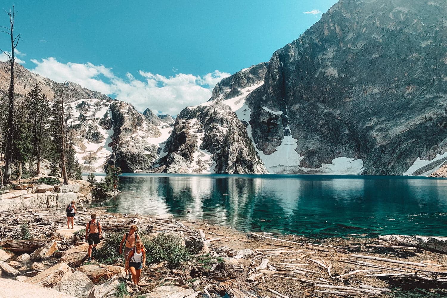 hikers walking along a rocky shore by a vibrant blue lake surrounded by mountains.