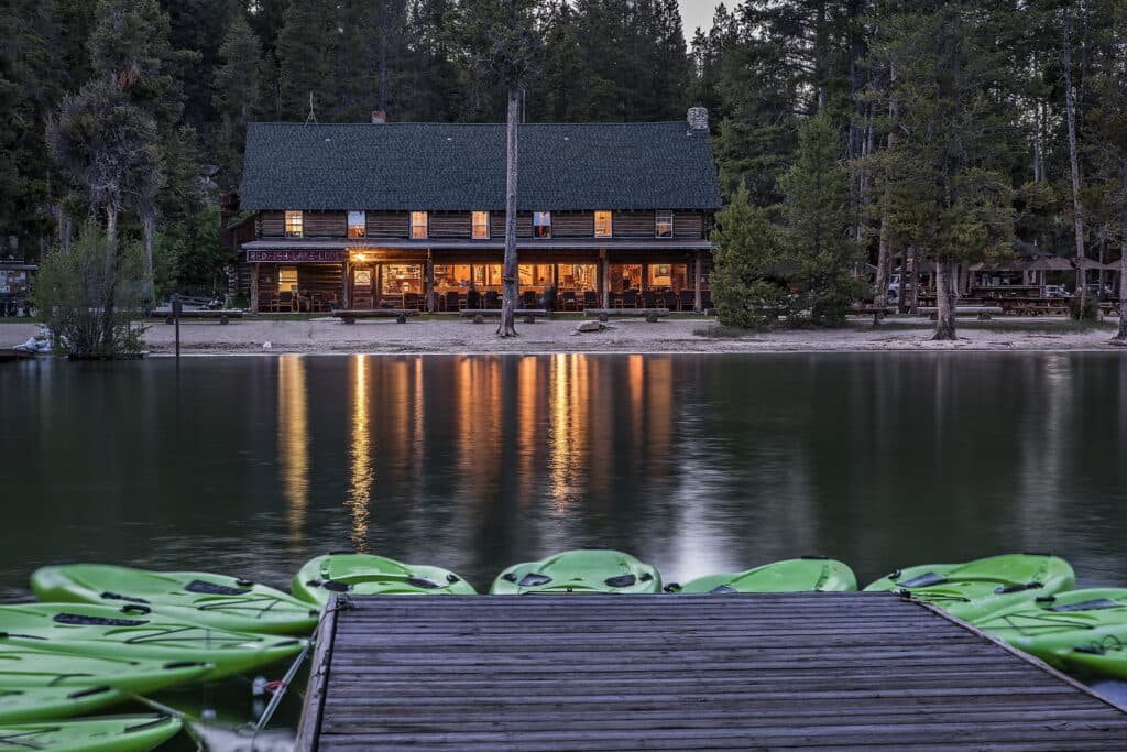 A rustic lodge with a green roof is reflected in a calm lake at dusk, with kayaks lined up on the dock in the foreground.