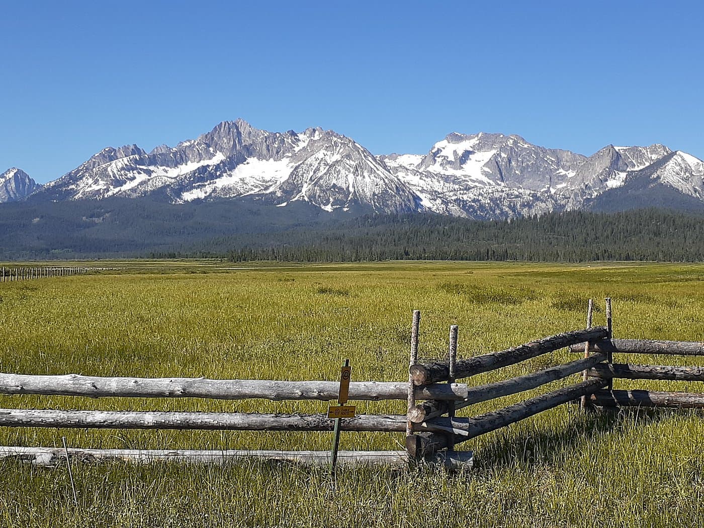 A scenic view of snow-capped mountains behind a grassy meadow with a wooden fence in the foreground.