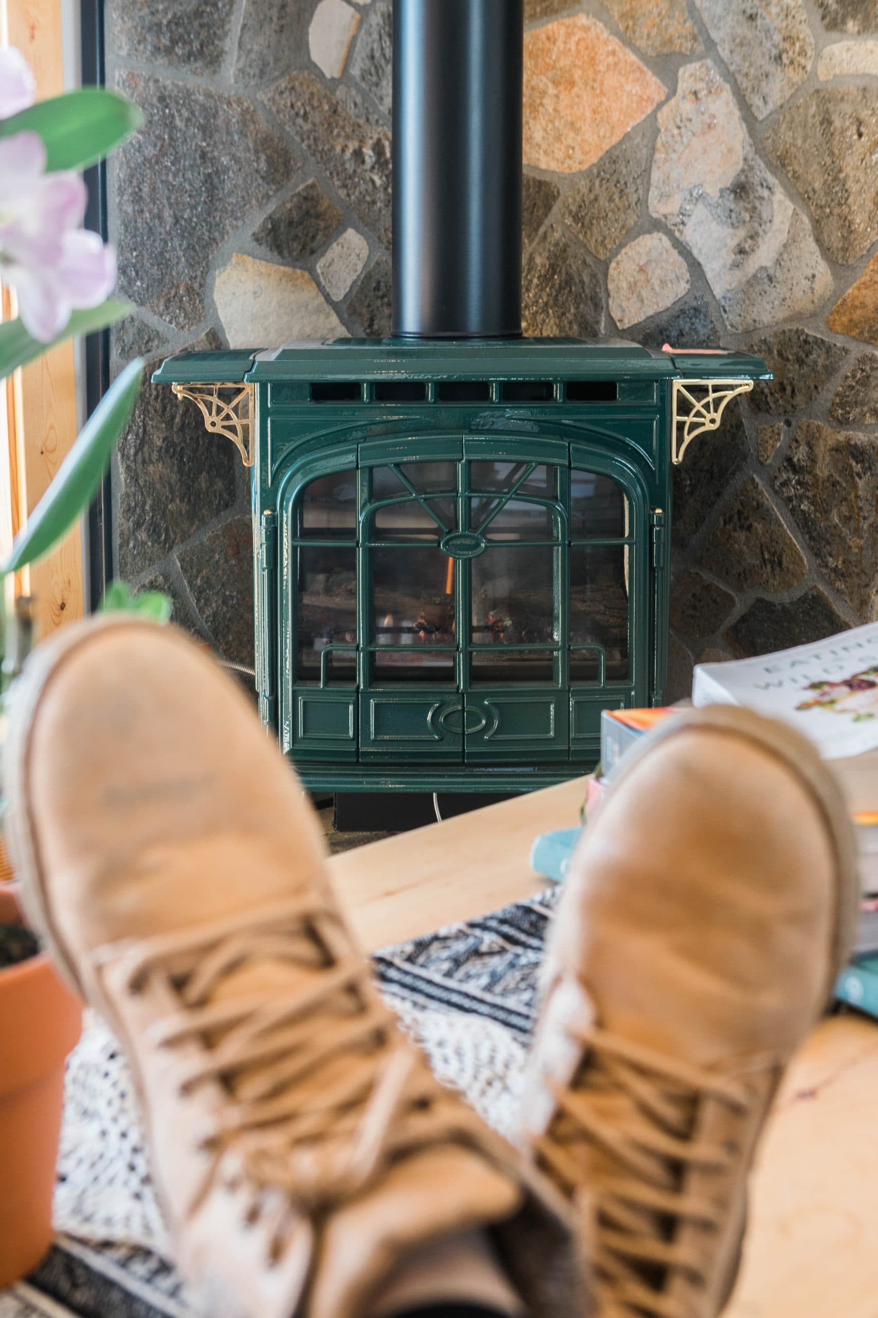 A pair of brown shoes rests on a rug in front of a green wood stove against a stone wall.