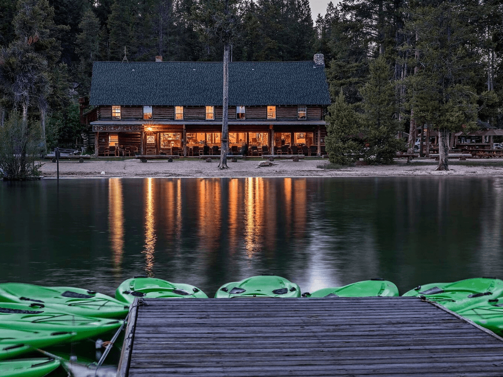 A rustic lodge by a calm lake at dusk, with green kayaks moored at a wooden dock.