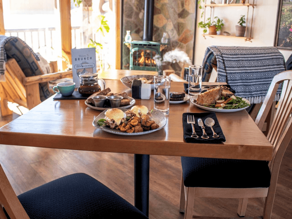 A beautifully set wooden table with brunch dishes, drinks, and a cozy fireplace in the background.