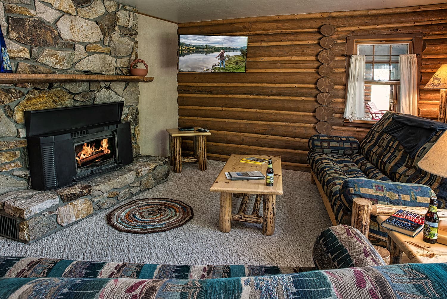 Cozy living room with a stone fireplace, rustic furniture, and a TV mounted on a log wall.