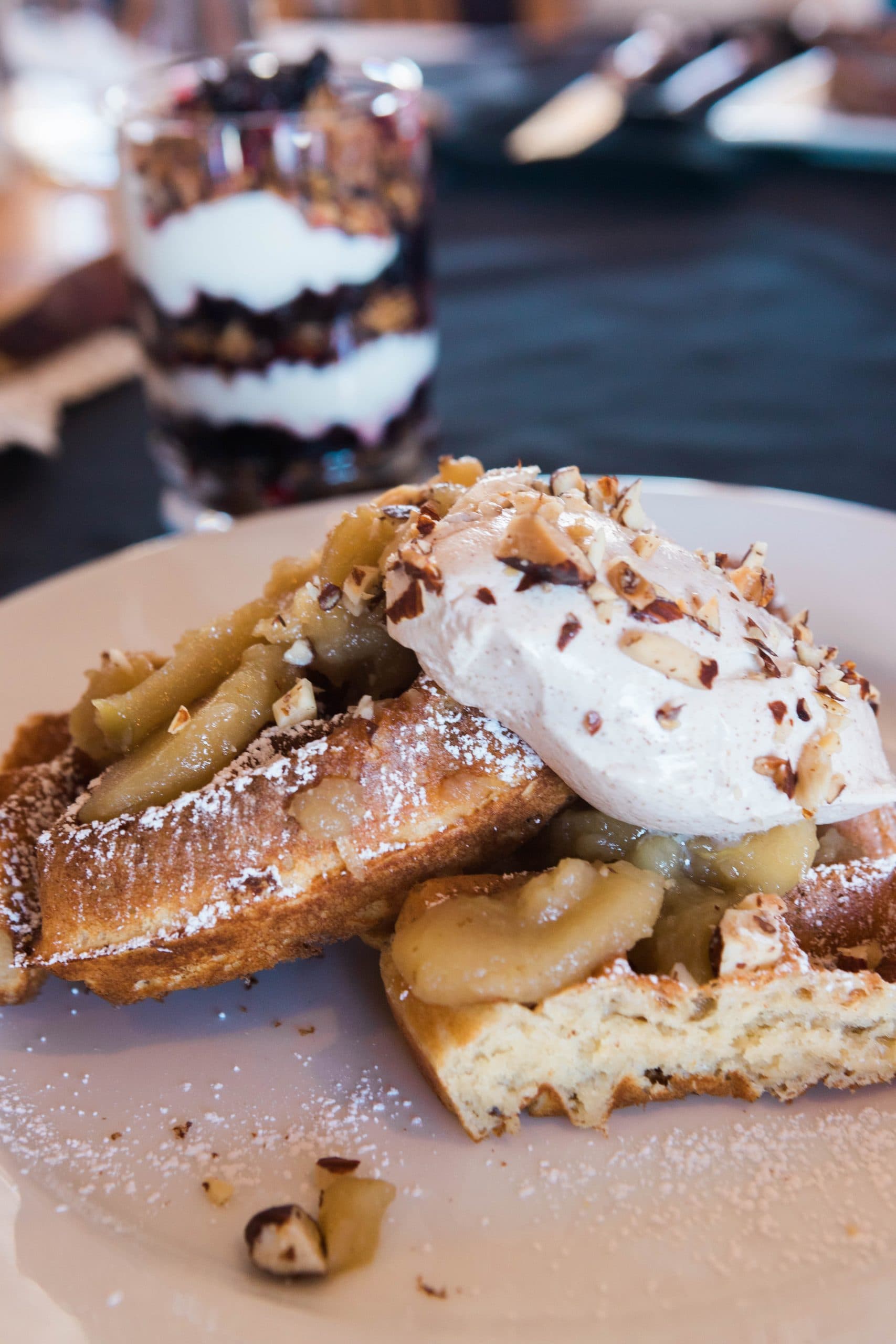 A plate featuring two waffles topped with apple sauce, whipped cream, and chopped nuts, alongside a layered dessert in the background.