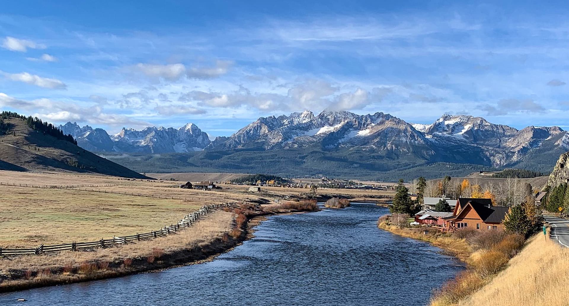 A tranquil river flows through a valley with snow-capped mountains in the background and a cabin nearby.