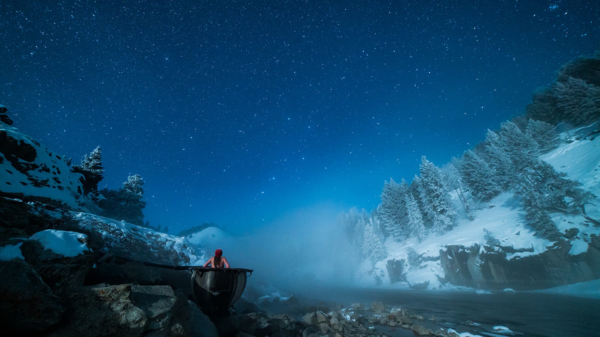 A person in a red jacket sits on a wooden boat by a snowy river under a starry night sky.