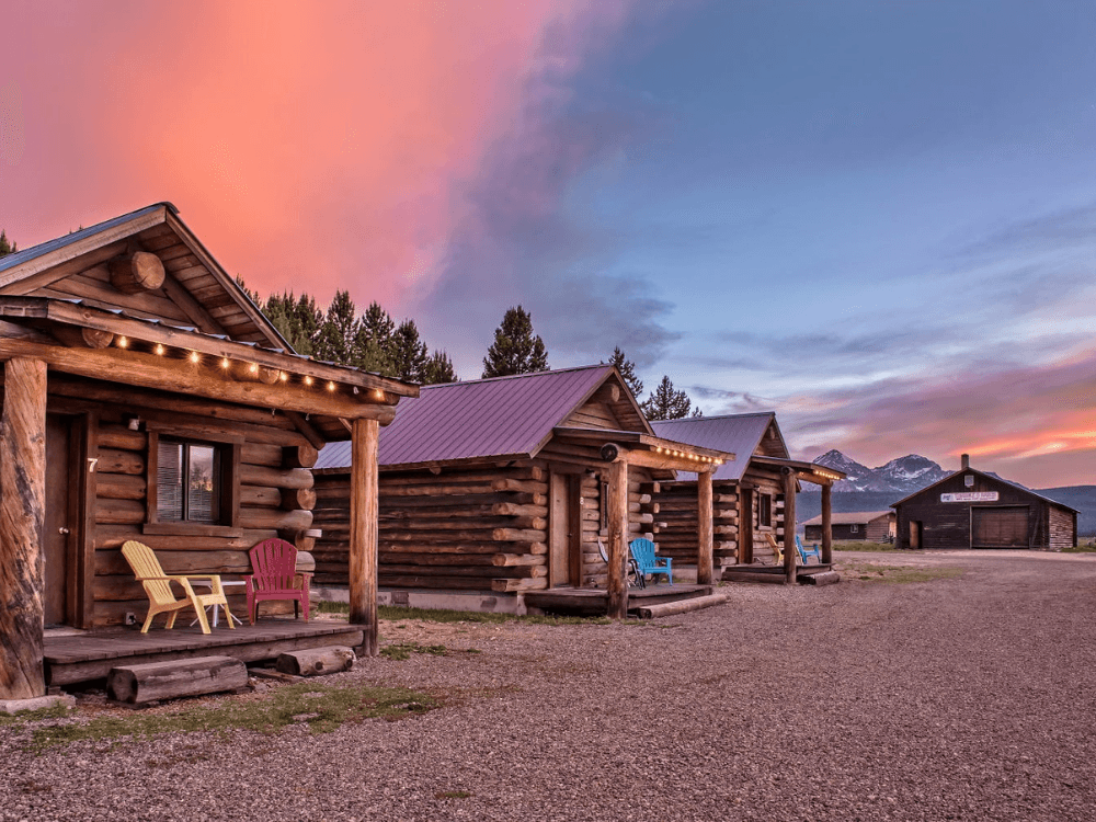 A row of rustic log cabins with colorful chairs at sunset, surrounded by mountains and trees.