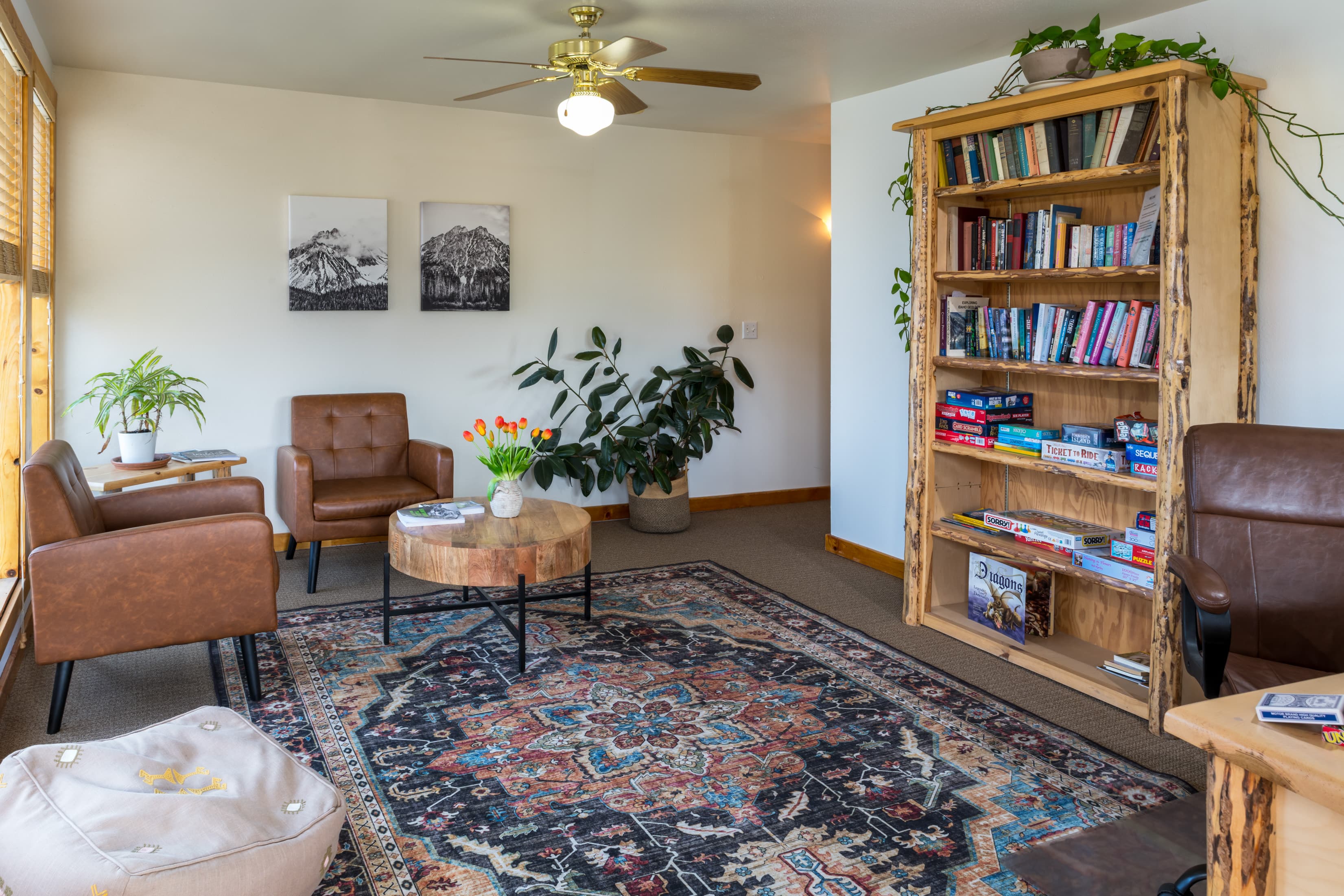 A cozy living room featuring two brown chairs, a wooden bookshelf filled with books, a circular coffee table, and plants, all set on a patterned rug.