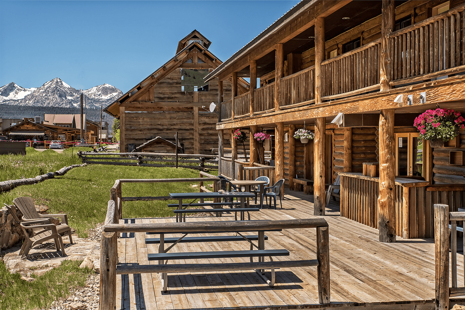 A wooden porch area in front of log cabins with colorful flower baskets, set against a backdrop of mountains.