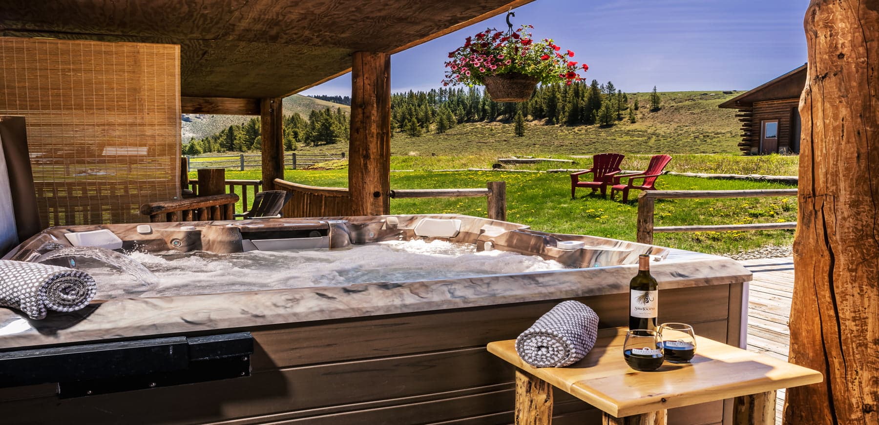 A hot tub with towels and wine glasses overlooks a scenic green landscape and wooden chairs.