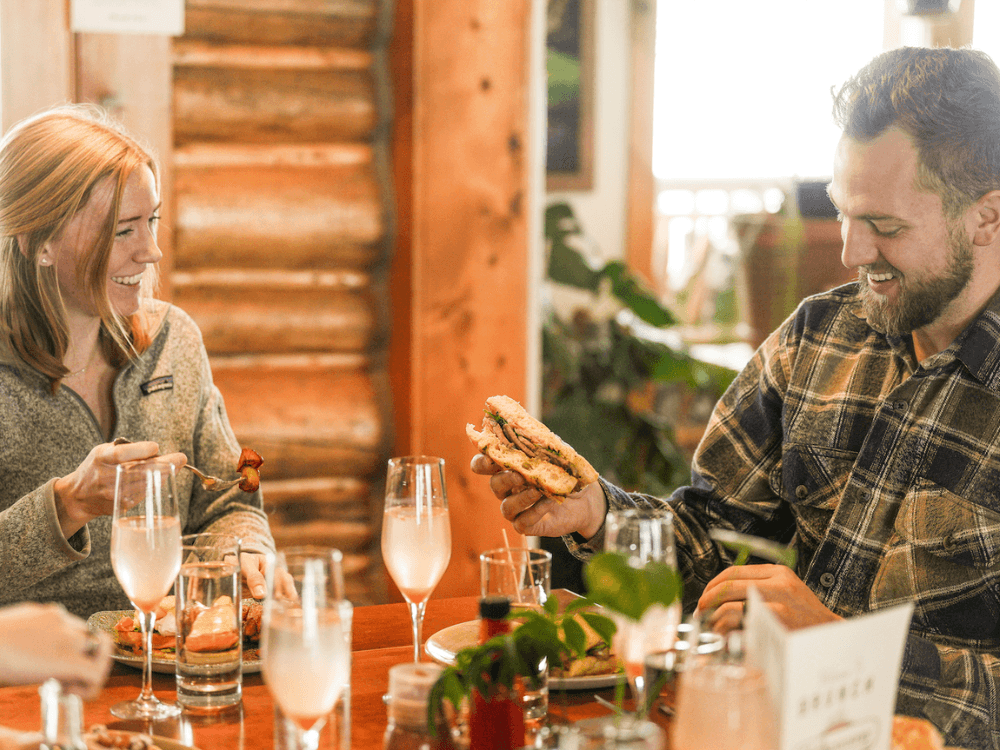A woman and a man enjoy a meal together at a wooden table, smiling and sharing food in a cozy setting.