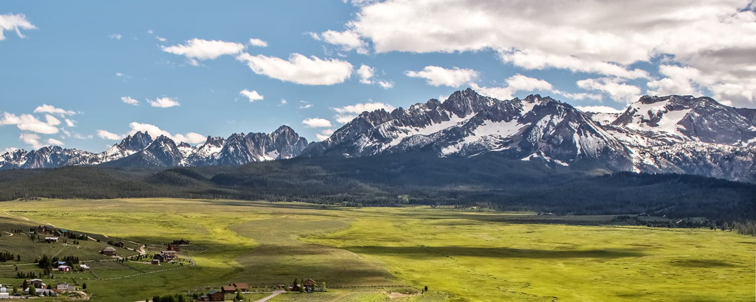 A panoramic view of snow-capped mountains rising over a green valley under a partly cloudy sky.