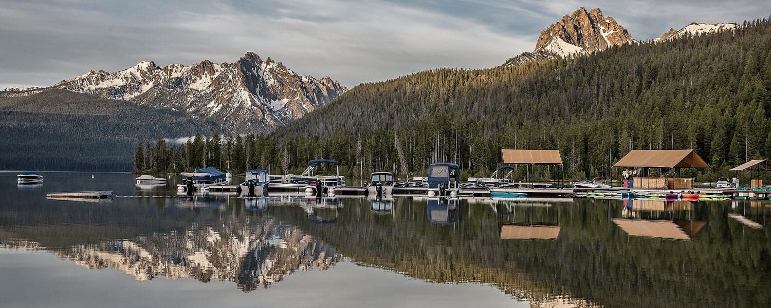 A serene lake scene with boats and a tree-lined shore, reflecting rugged mountains in the background.