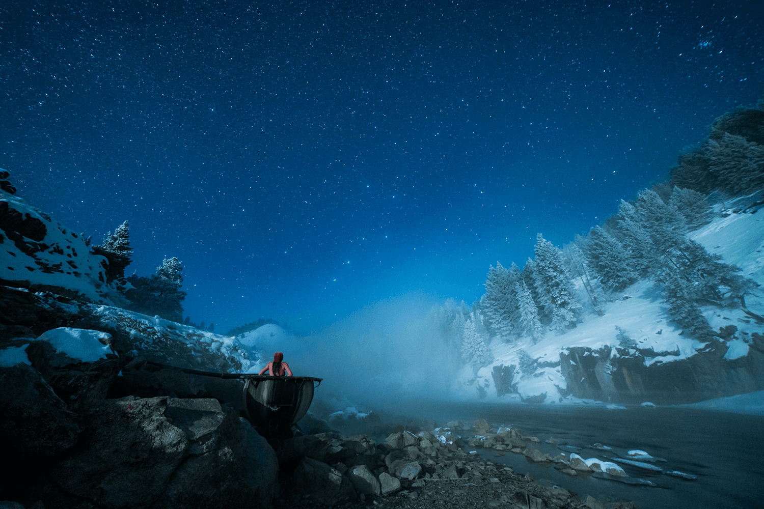 A person in a red jacket sits on a rock by a snowy river under a starry night sky.